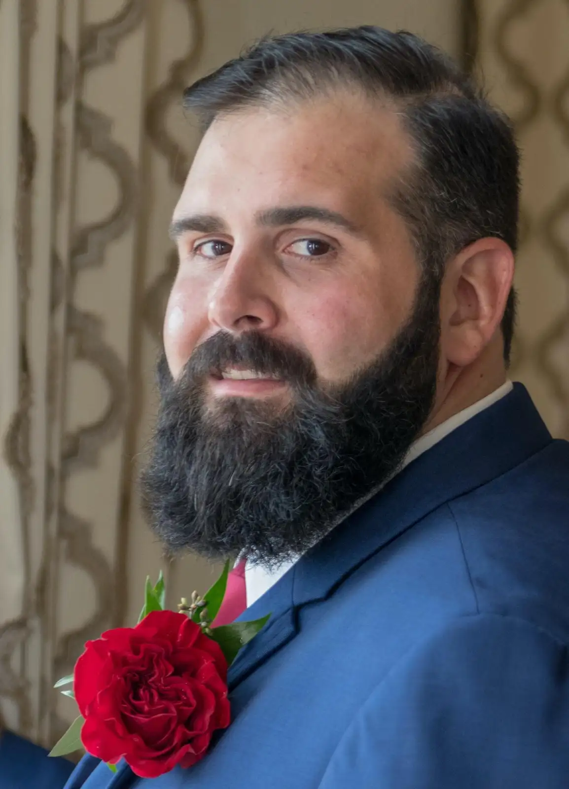 A man with a full dark beard, wearing a blue suit and a red rose boutonniere, looks directly at the camera with a subtle…