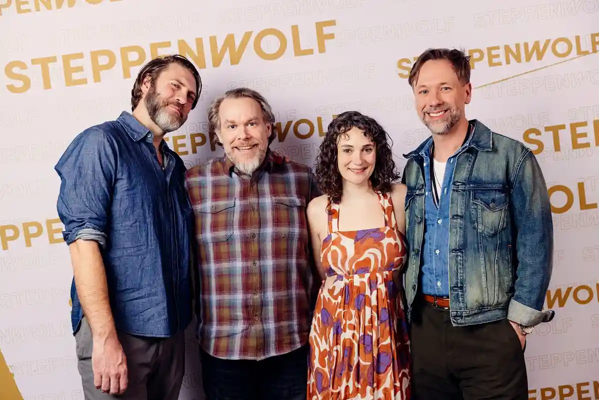 Four people stand together in front of a backdrop repeatedly printed with the word 'STEPPENWOLF' in large gold letters. The…