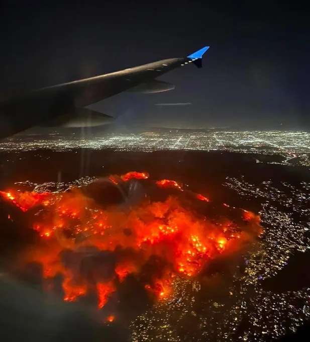 An aerial photo taken from an airplane window shows the wing in the upper left, with a sprawling cityscape below illuminated…