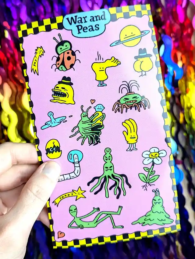 A hand holds a pink card with a black-and-yellow checkered border, featuring a collection of whimsical, cartoonish alien and…