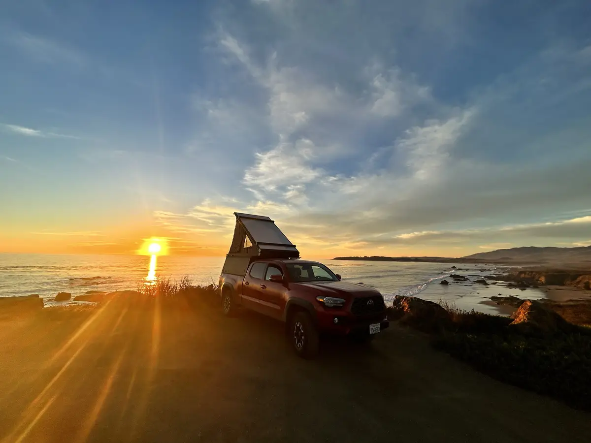 A red pickup truck with a rooftop tent is parked on a coastal road at sunset, with the sun setting over the ocean and hills…