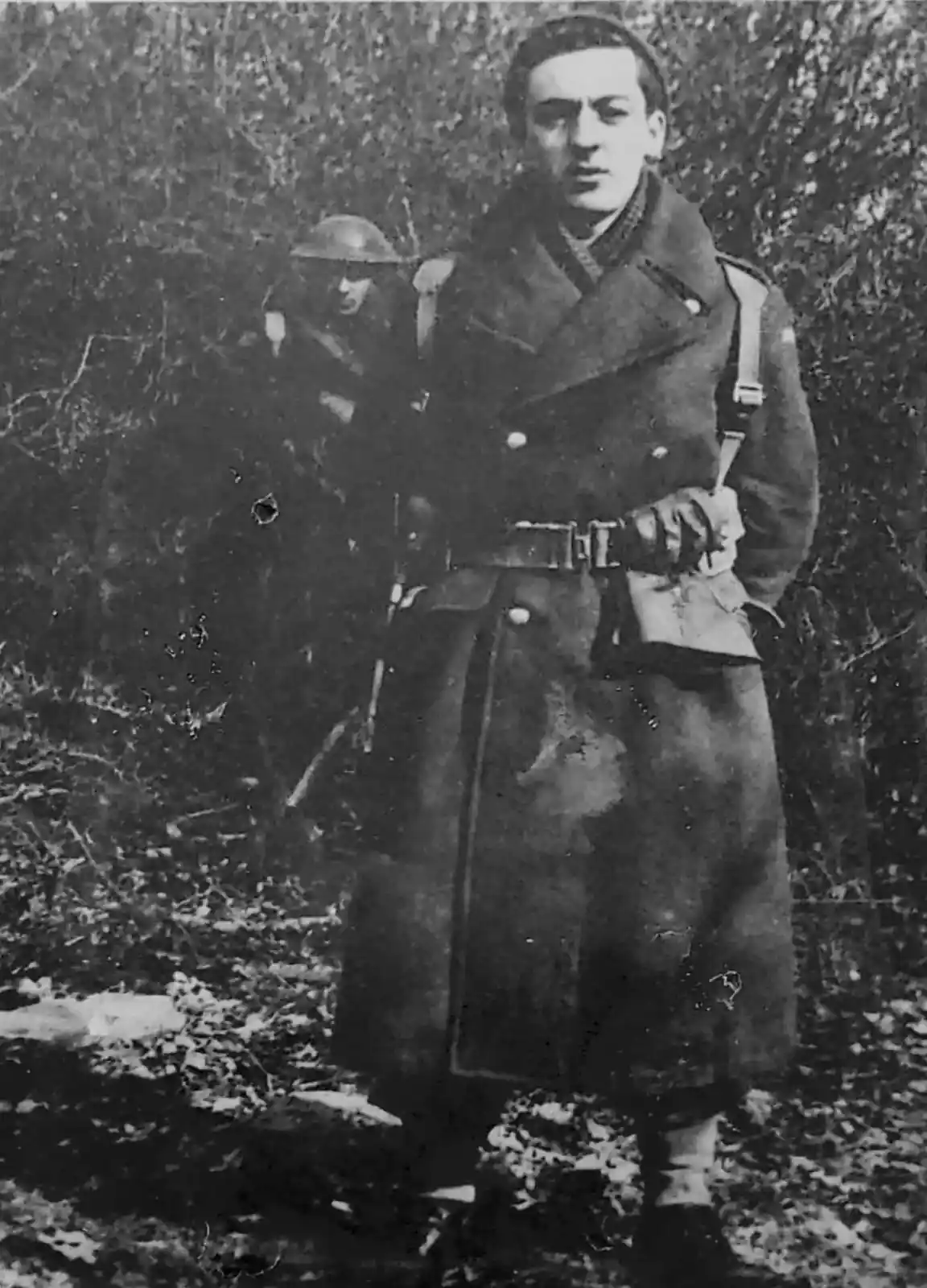A grainy black-and-white photo shows a young man in a long military coat and gloves standing outdoors, with another soldier…