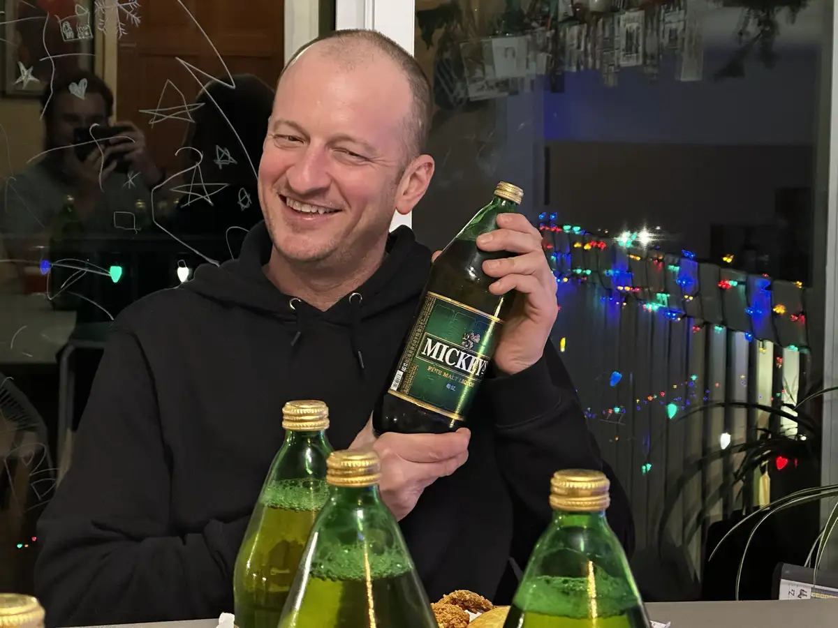 A man in a black hoodie smiles while holding a large green bottle labeled 'MICKIES FINE MALT LIQUOR', with several smaller…