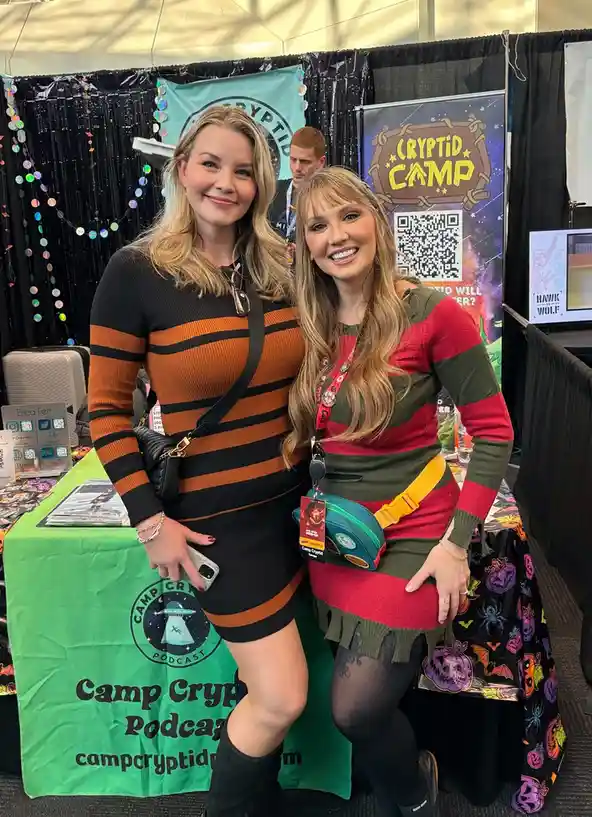 Two women pose together at a 'Camp Cryptid' event booth, both wearing striped dresses. The woman on the left wears a black…
