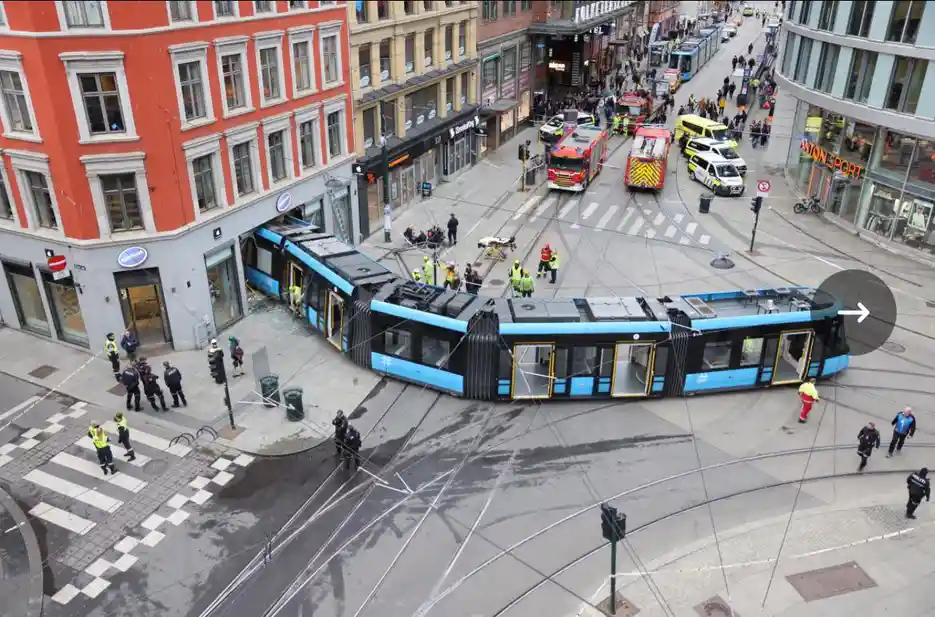 An aerial view shows a blue and black tram that has crashed into a building and is bent into a sharp, unnatural curve, with…