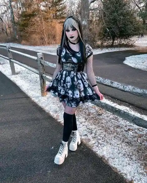 A person with gothic styling, including black lipstick, fishnet sleeves, and a dress covered in printed ghostly faces…