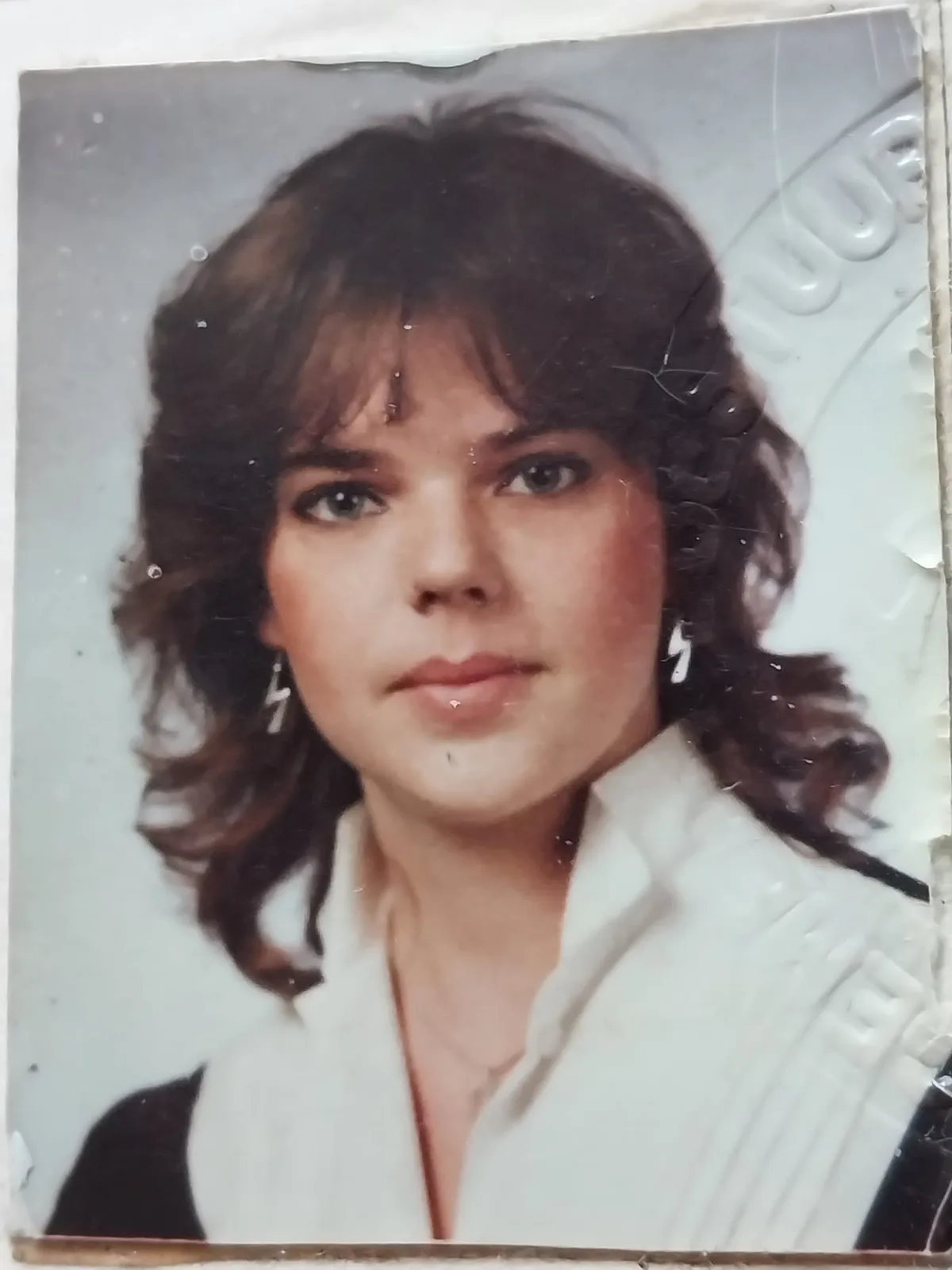 A visibly aged, slightly damaged studio portrait of a young woman with voluminous 80s-style brown hair, wearing a white…