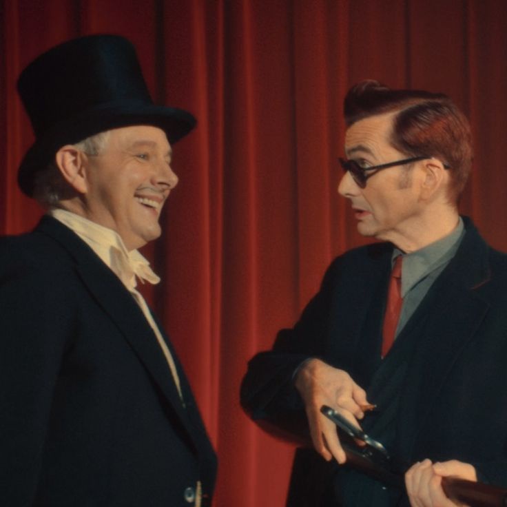 Two men in suits stand before a red curtain; one wears a top hat and laughs while the other, in glasses and holding a…