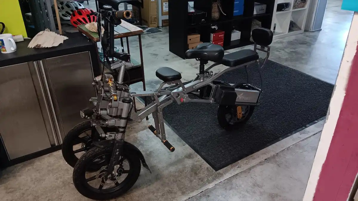 The image shows a silver Freeda electric scooter with an unusually complex and exposed mechanical design, featuring multiple…