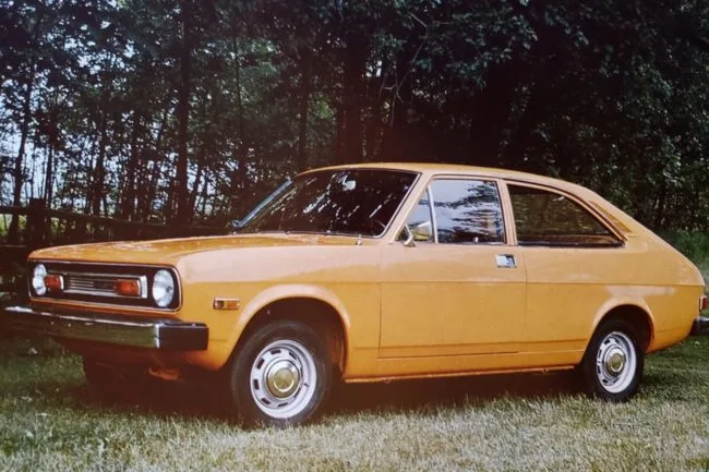 A vintage mustard-yellow two-door coupe is parked on grass with a forest background. The car has a boxy, retro design with…
