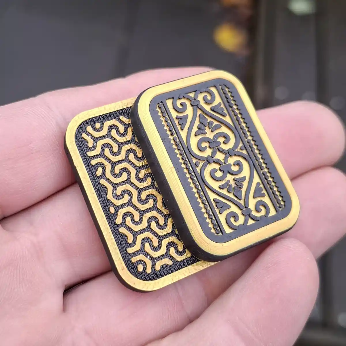 A hand holds two small, rectangular, 3D-printed objects resembling playing cards with ornate black and gold patterns. One…