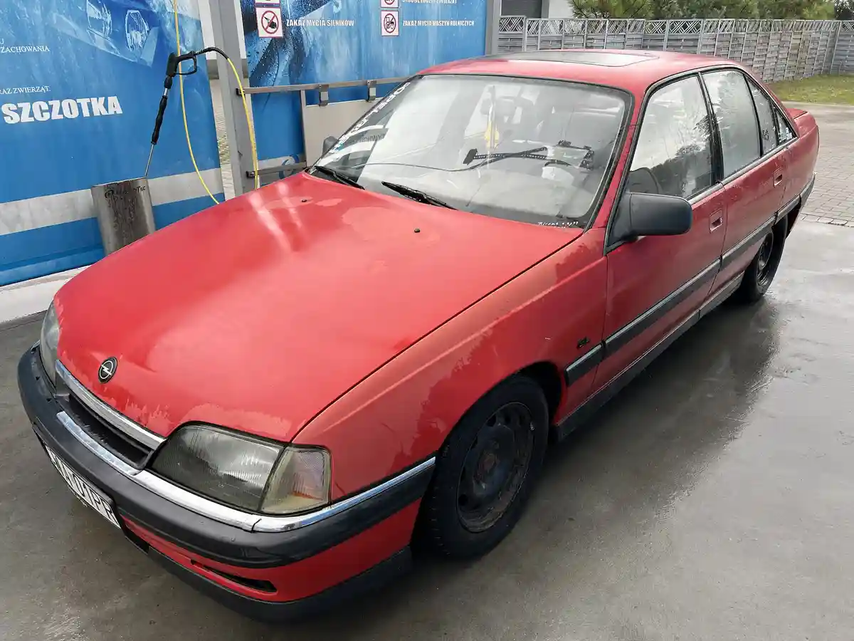 A red Opel sedan is parked at a car wash, its hood and body showing signs of dirt and wear, with a yellow hose and nozzle…