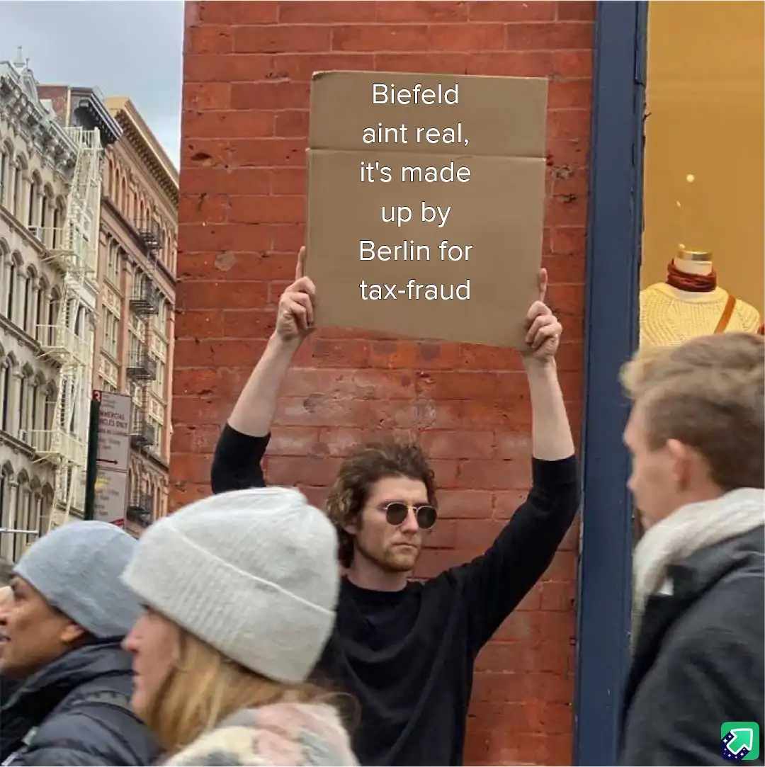A man with curly hair and sunglasses holds up a cardboard sign in front of a brick wall on a city street. The sign reads…