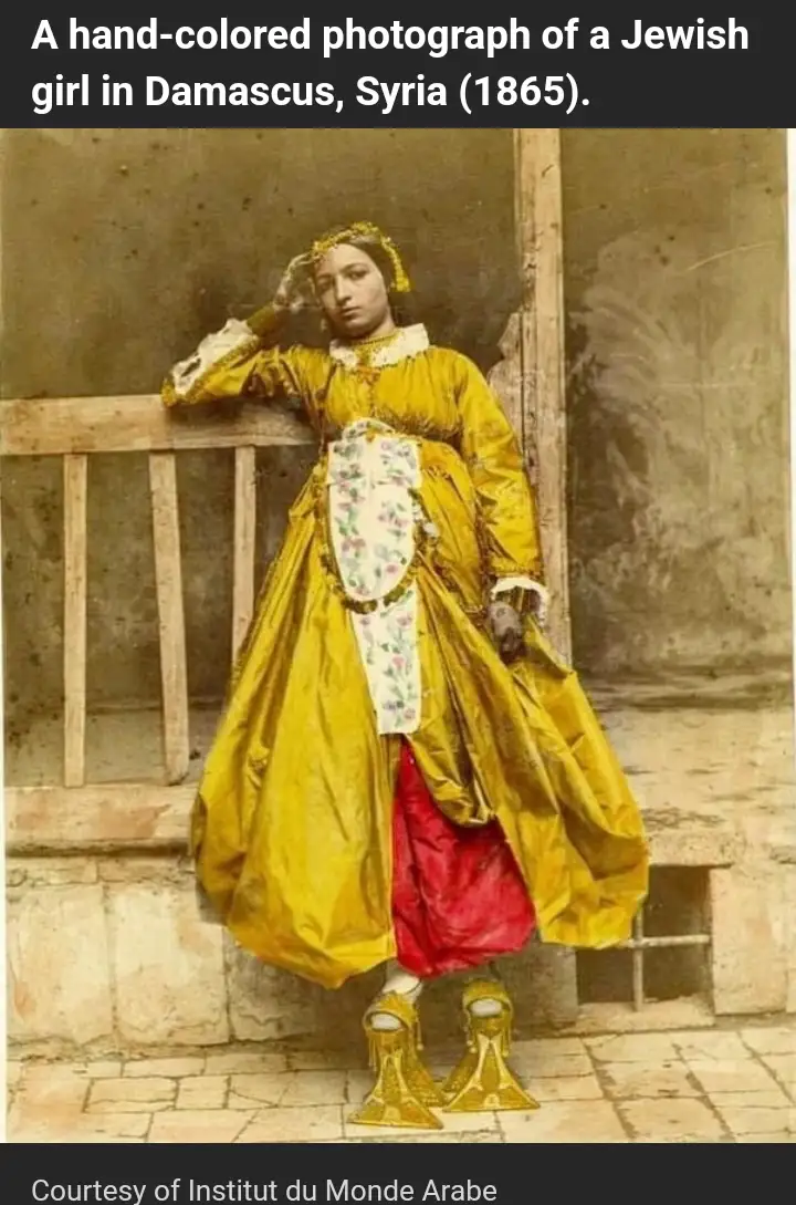 A hand-colored 1865 photograph shows a young woman in ornate yellow and red traditional dress, standing beside a wooden…