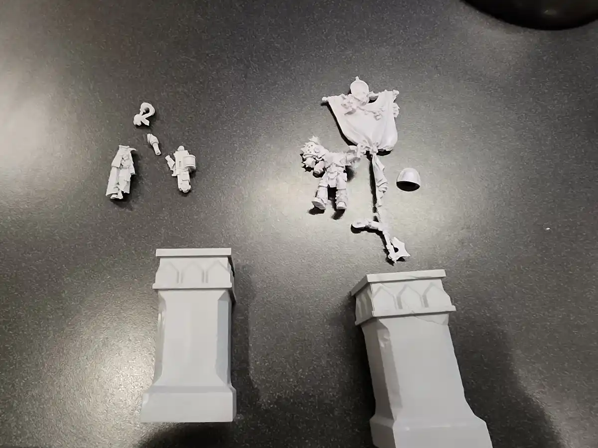 Two white, unassembled Warhammer 40k miniature kits lie on a gray surface, each with scattered parts and bases. The text…