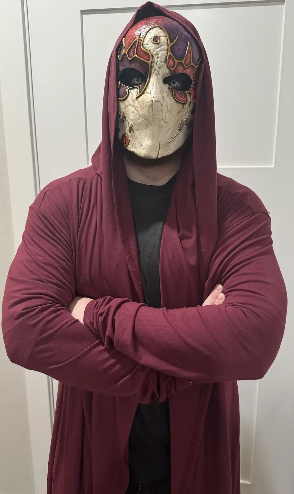 A person wearing a maroon hooded robe and a detailed, ornate mask with red, purple, and gold designs stands with arms…