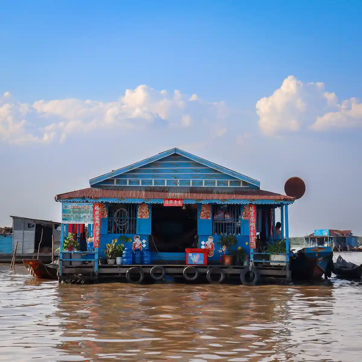 A vibrant blue floating house on murky water under a partly cloudy sky, decorated with red Chinese New Year banners, potted…