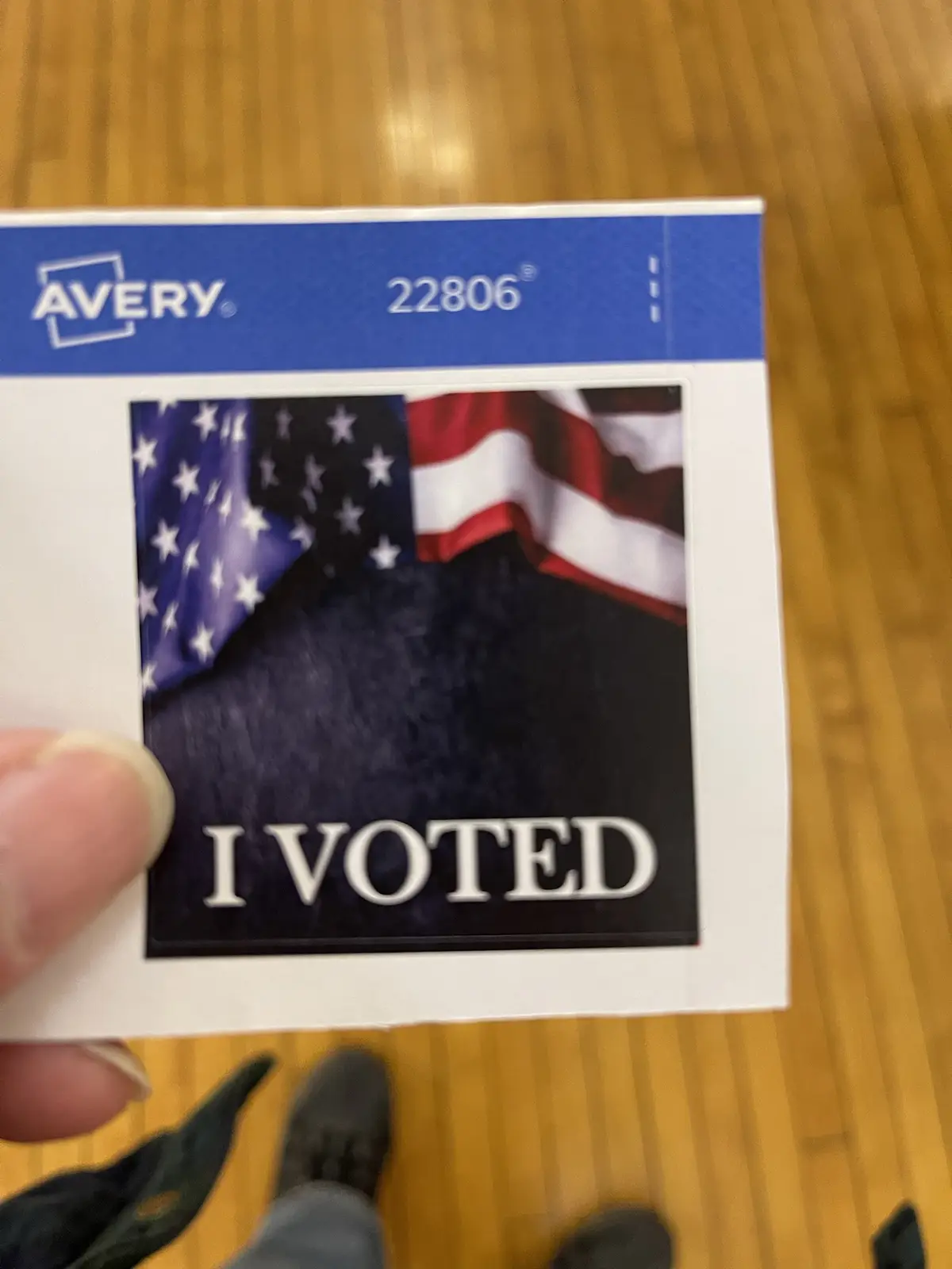A person holds up a standard Avery brand 'I VOTED' sticker, which features an American flag and the text 'I VOTED' against a…