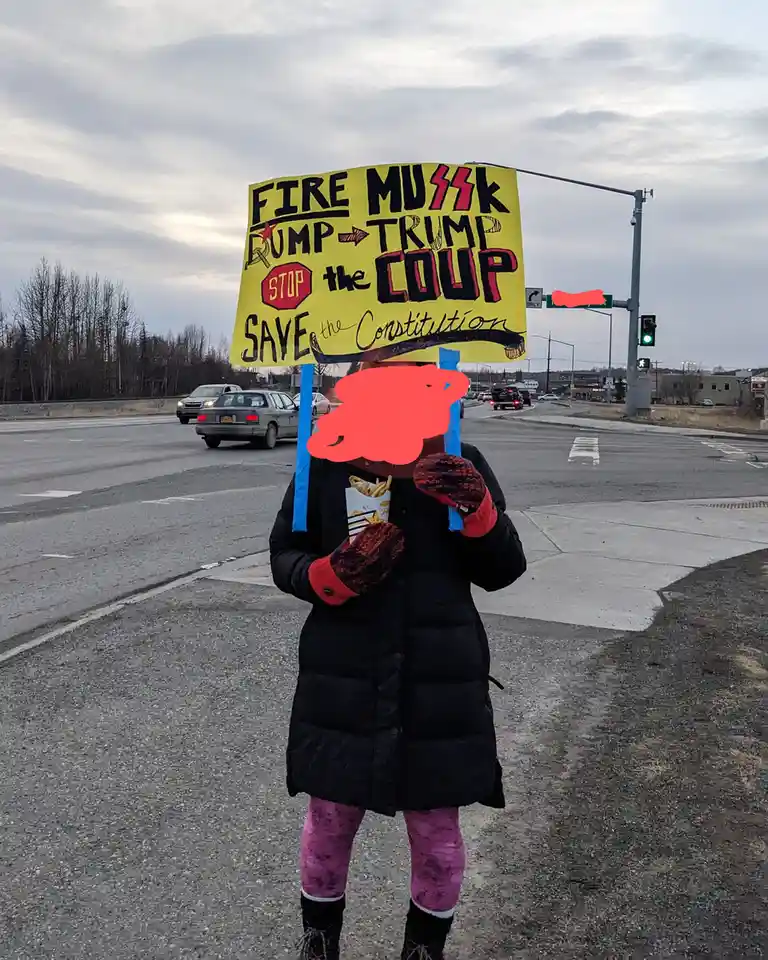 A person wearing a black puffer coat, pink leggings, and patterned mittens stands at a street corner holding a large yellow…