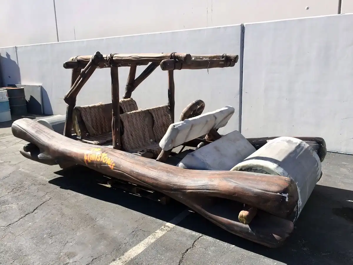 A weathered, rustic vehicle resembling a Flintstones car, constructed from logs and stone-like materials, sits parked…