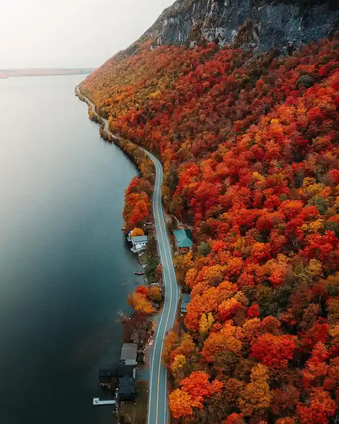 An aerial photo shows a winding road hugging a lake, bordered by a dense forest ablaze with vibrant red, orange, and yellow…