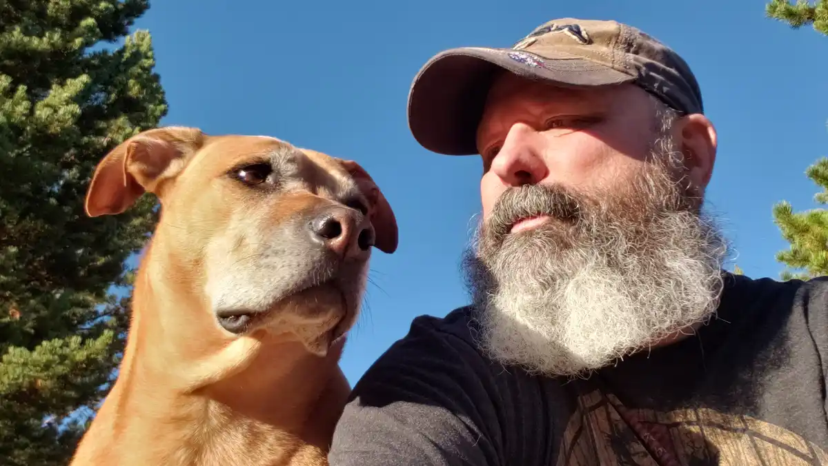 A low-angle selfie shows a man with a long gray beard and a baseball cap standing beside a tan dog, both looking off to the…