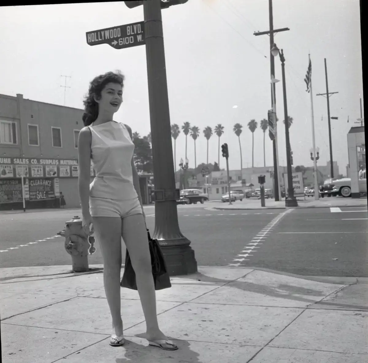 A vintage black-and-white photo shows a smiling woman in a white sleeveless top and shorts standing on a sidewalk near a…