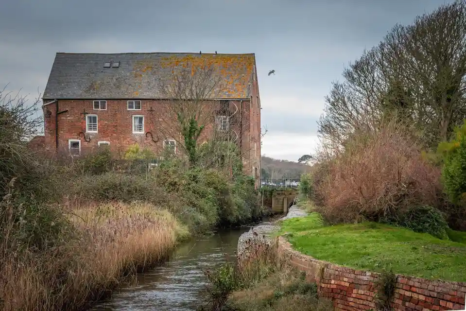A red brick building with a mossy roof stands beside a narrow canal, surrounded by overgrown bushes and bare trees under an…