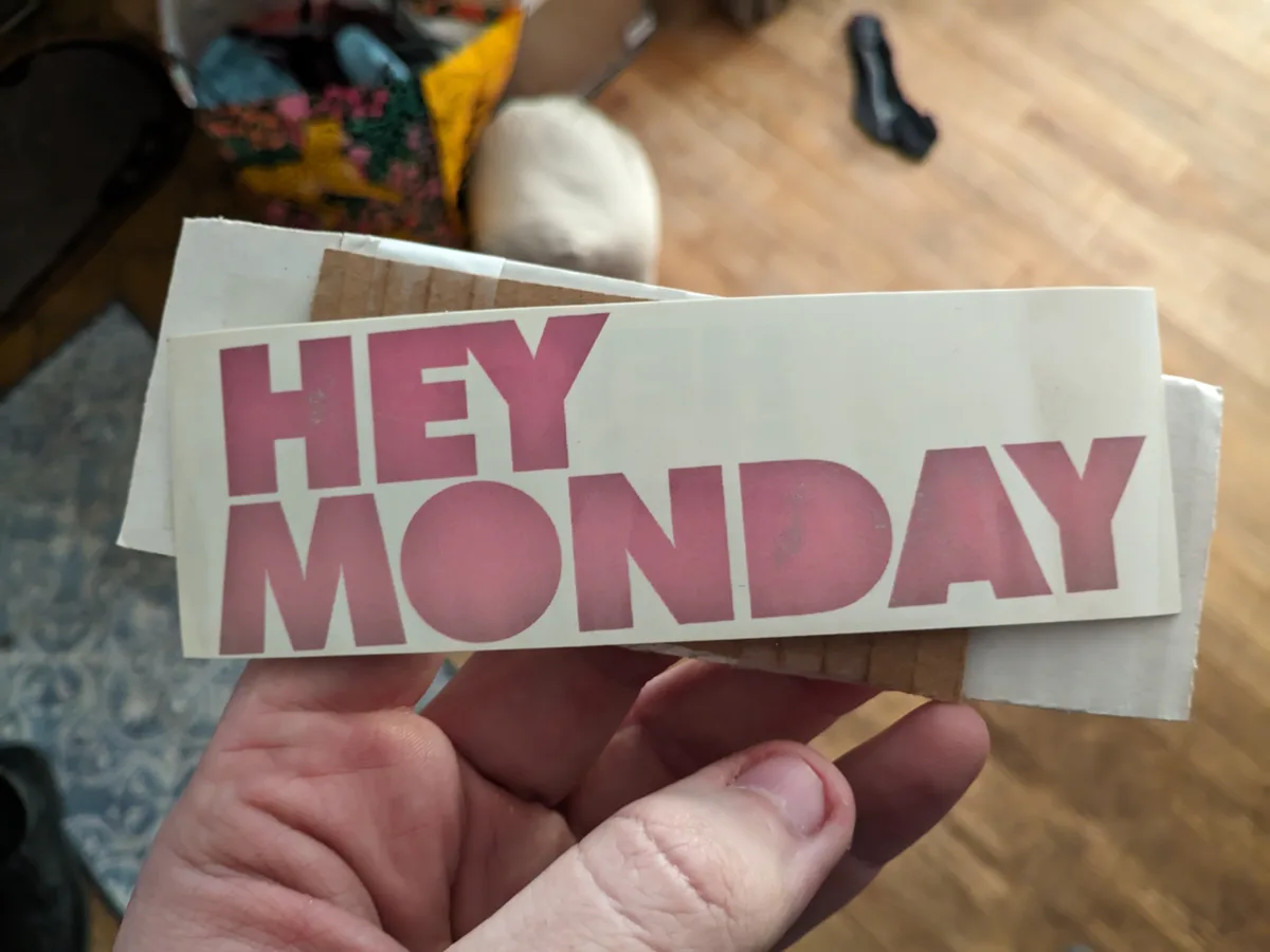 A hand holds a worn, pink-lettered sticker that reads 'HEY MONDAY' against a blurry background of a wooden floor and…