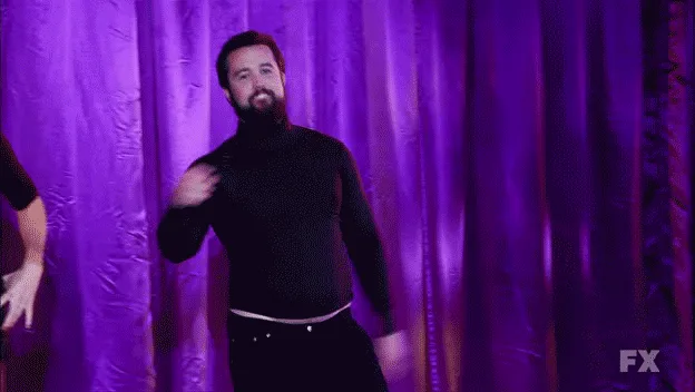 A man in a black turtleneck dances in front of a purple curtain, with a woman appearing behind him in some frames. The…
