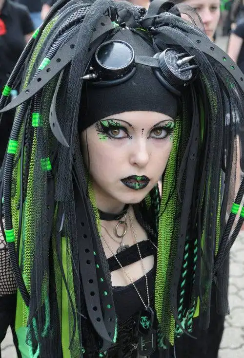 A person with elaborate black and neon green hair made of tubes and straps, wearing goggles on their head, heavy makeup…