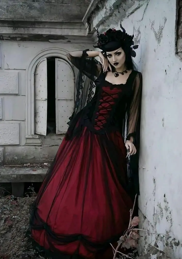 A woman in a gothic red and black dress with lace, holding a gun, leans against a crumbling white wall next to an arched…