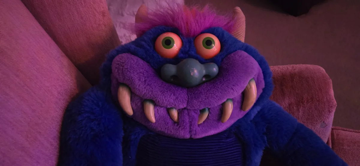 A plush toy resembling a blue and purple monster with large green eyes, sharp teeth, and horns sits on a pink couch. The…