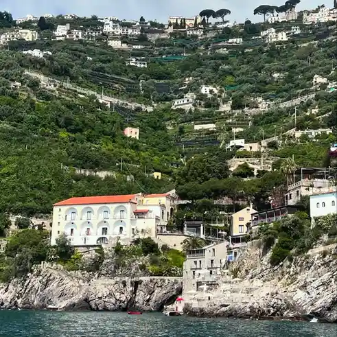 A scenic photo of a coastal hillside town with white buildings, terraced gardens, and a rocky shoreline overlooking the sea…