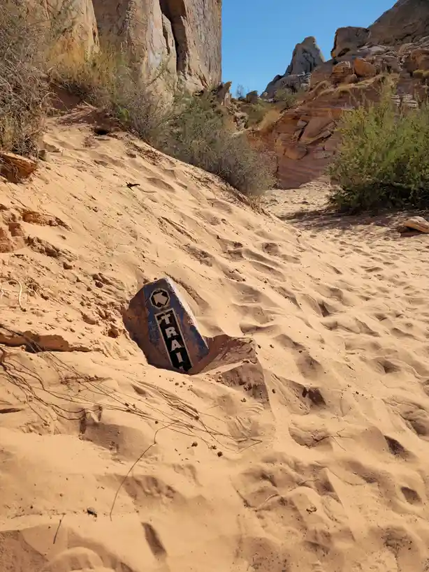 A weathered blue and white trail sign is half-buried in sand on a desert trail, with rocky cliffs and sparse vegetation in…