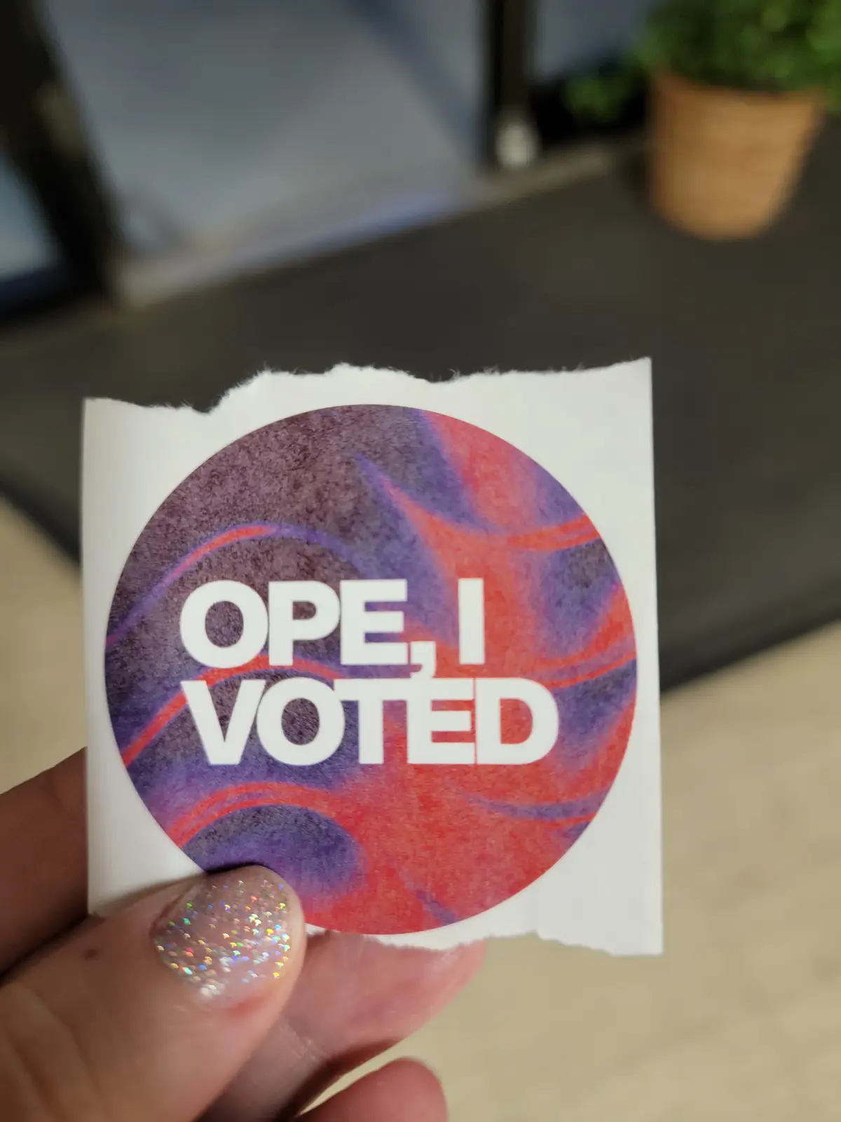 A hand holds up a circular sticker with a red and purple marbled background, featuring the text 'OPE, I VOTED' in bold white…