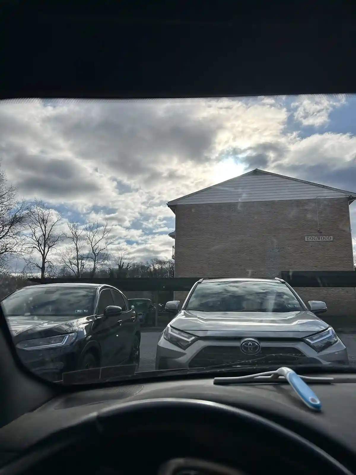A photo taken from inside a car shows a silver Toyota SUV parked next to a black SUV in a lot, with a brick building labeled…