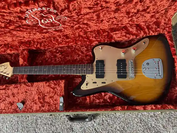 A Fender Jazzmaster electric guitar with a sunburst finish rests inside its open, plush red-lined case, which features an…
