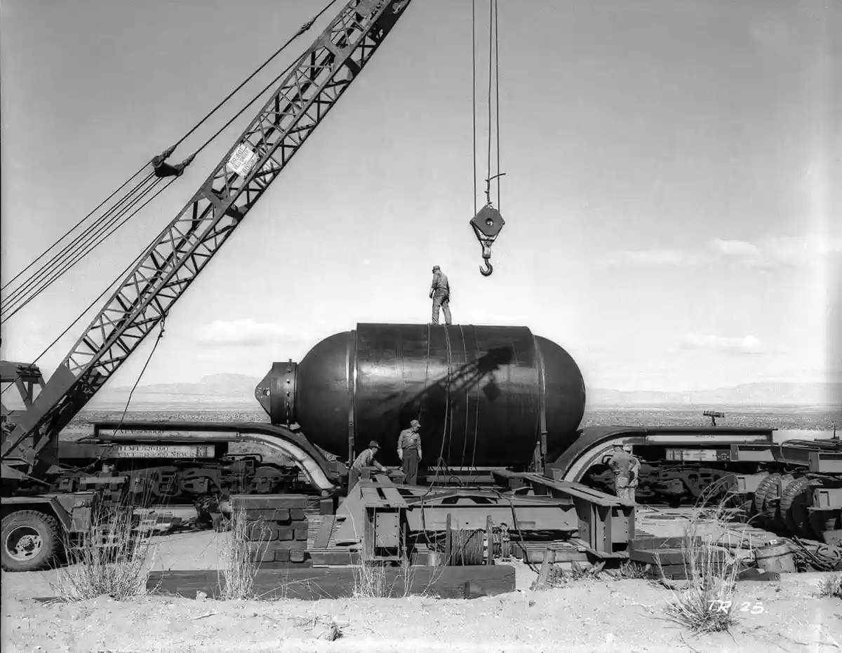 A black-and-white historical photo shows workers handling a massive, cylindrical object, likely a nuclear bomb, being lifted…