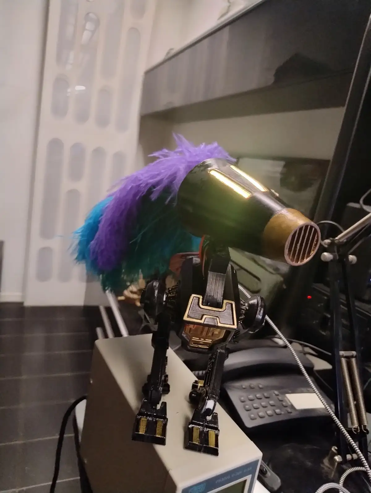 A small, black and gold robot figurine stands on a beige electronic device, its head replaced with a green hairdryer…