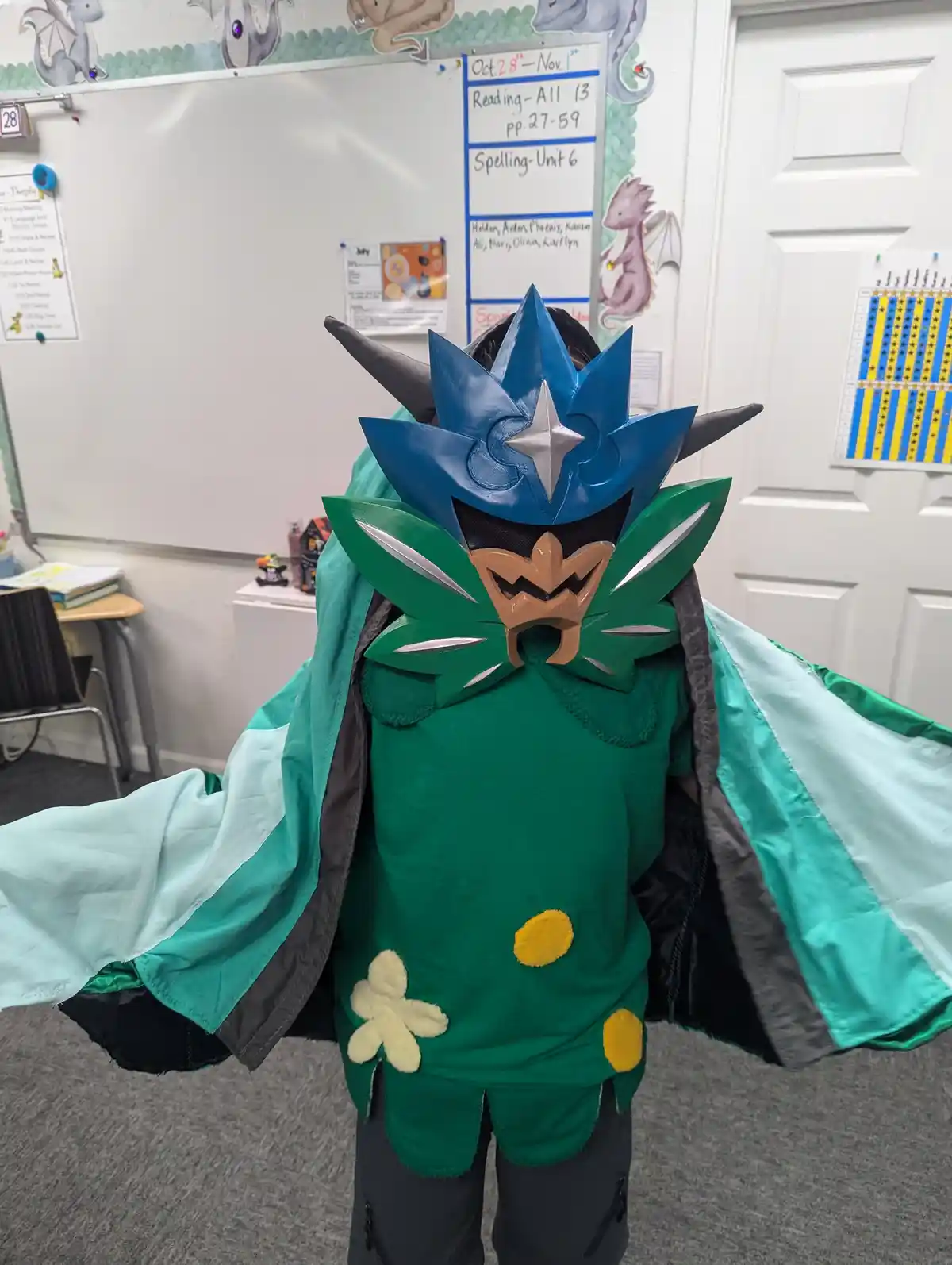 A child in a detailed cosplay of the Pokémon character Garchomp stands in a classroom, facing away from the camera, wearing…
