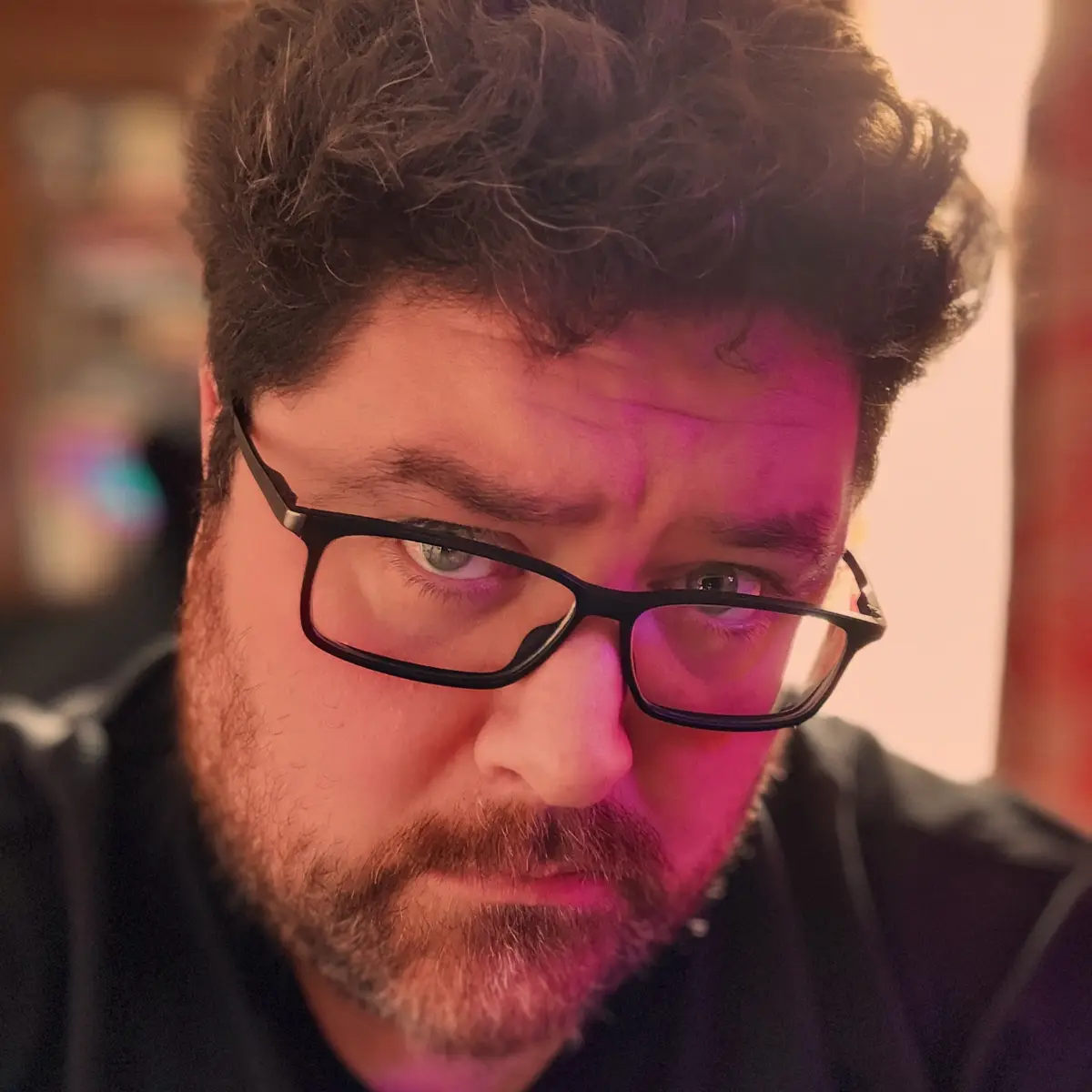 A close-up photo of a man with dark curly hair, a beard, and black-rimmed glasses, looking intensely at the camera with a…
