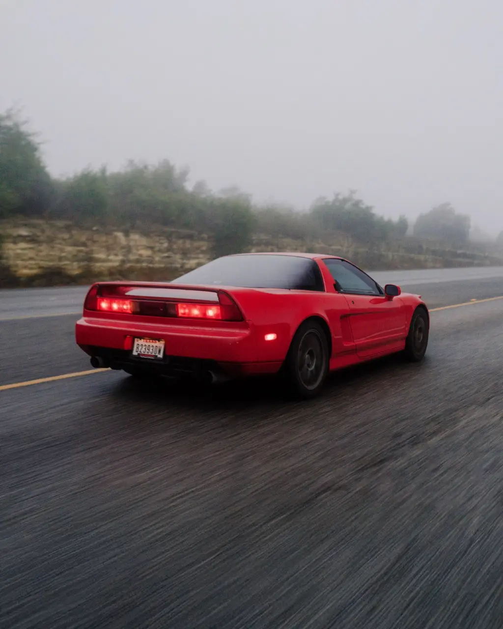A red sports car, likely an Acura NSX, is driving on a wet road through heavy fog. The car's taillights are illuminated, and…