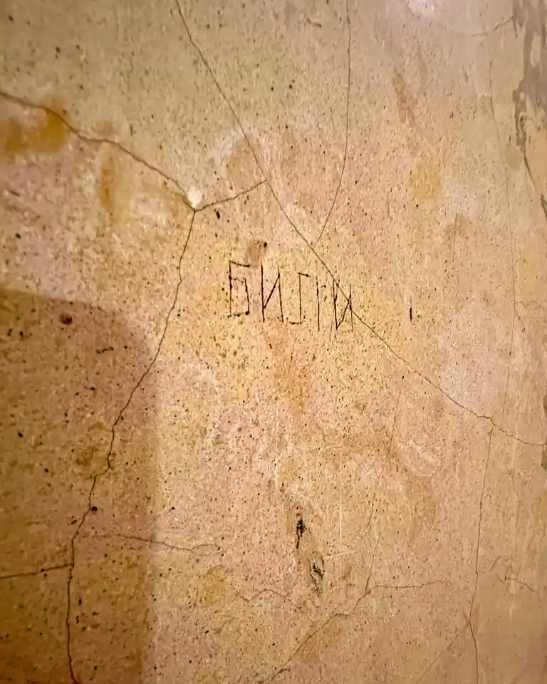 A close-up of a beige, cracked stone wall with faint, crudely scratched Cyrillic letters spelling 'билин' in the center. The…