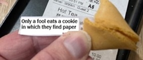 A close-up image shows a person's hand holding a golden-brown fortune cookie that has been cracked open, revealing a small…