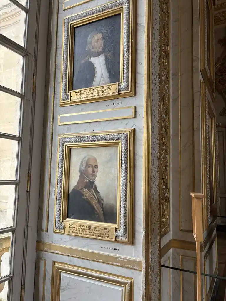 Two ornate, gold-framed historical portraits hang on a marble wall with gold trim, each with a plaque below detailing the…