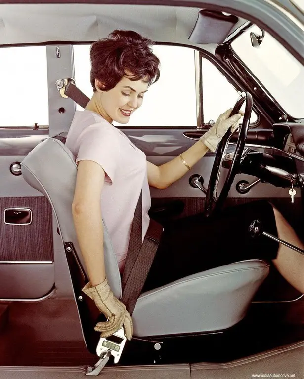 a vintage photo of a smiling woman in a 1950s car, wearing gloves and a pink dress, fastening her seatbelt with a modern…