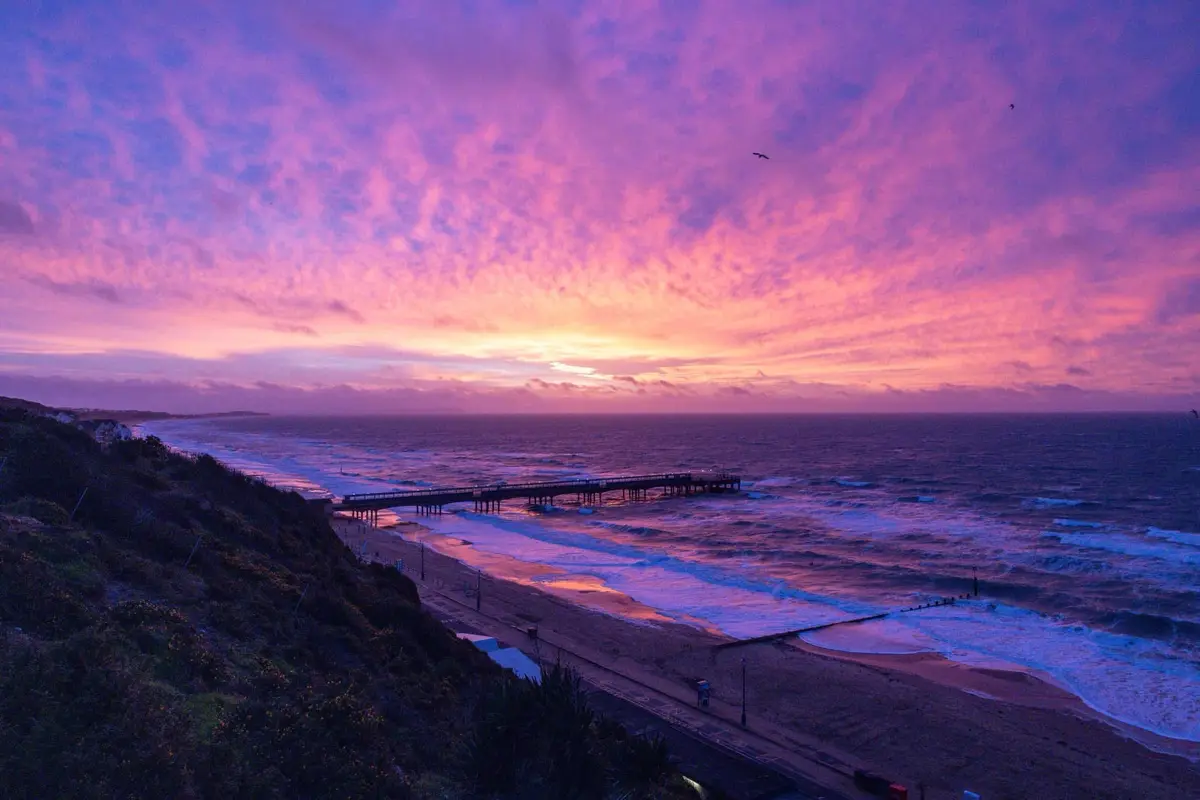A scenic photograph of a beach at sunset or sunrise, featuring a long pier extending into the ocean, waves crashing on the…