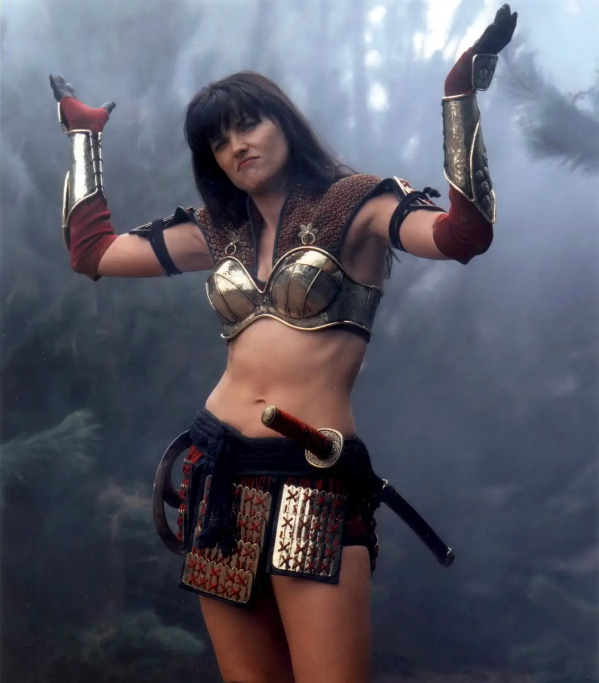 A woman in a fantasy warrior outfit with metallic arm guards, a gold bra-like chest piece, and a sword at her hip stands…