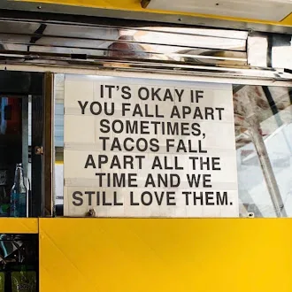 A sign inside a yellow taco truck or food cart reads: 'IT'S OKAY IF YOU FALL APART SOMETIMES, TACOS FALL APART ALL THE TIME…
